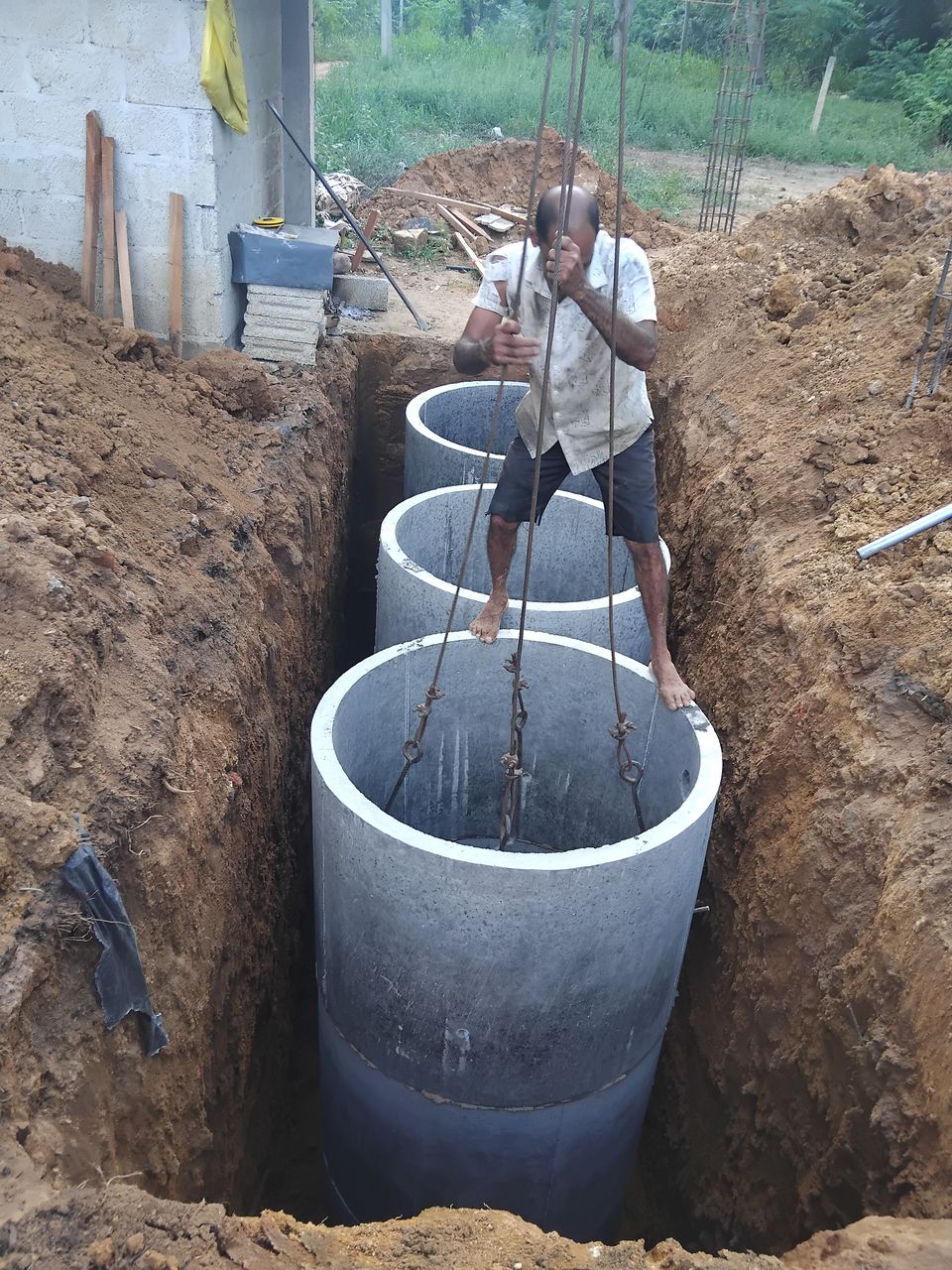 Concrete rings lowered on crane chains into a narrow trench beside a house being built, a worker guiding them into place