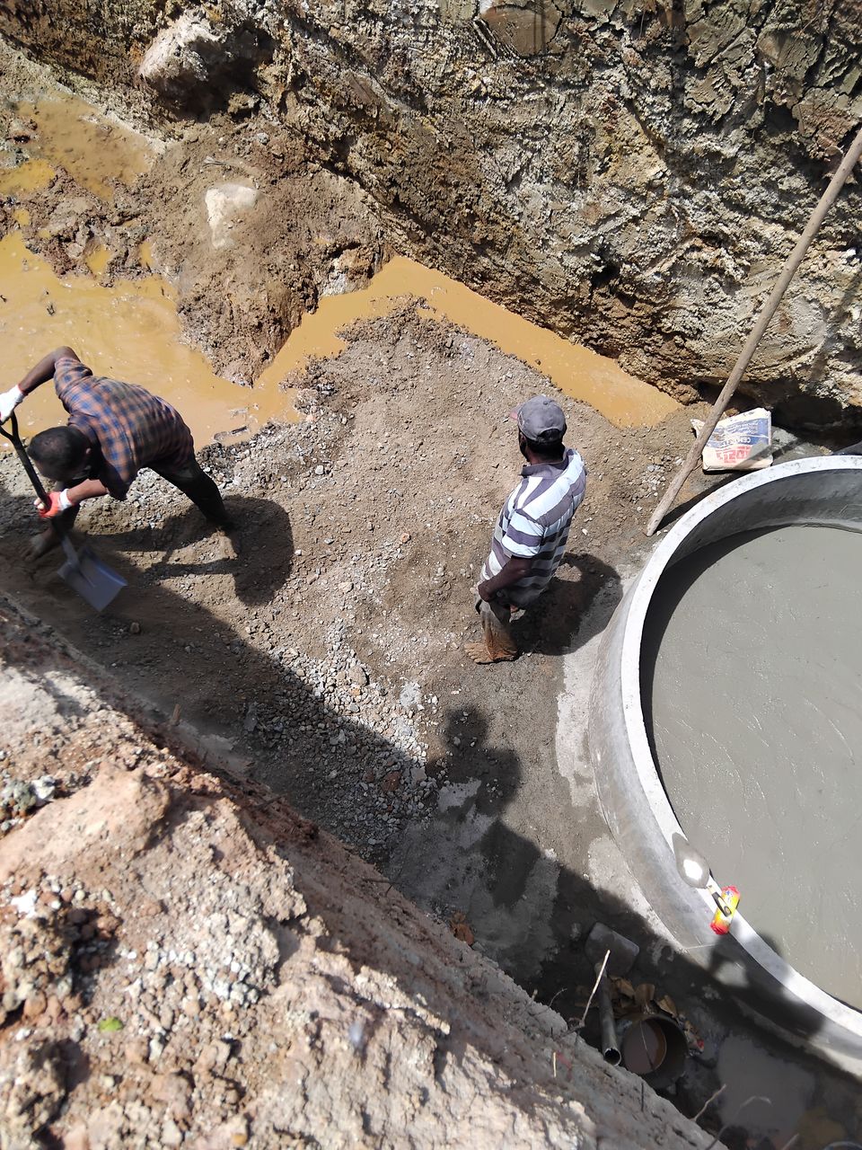 Workers spreading gravel at the bottom of an excavation beside a large concrete ring