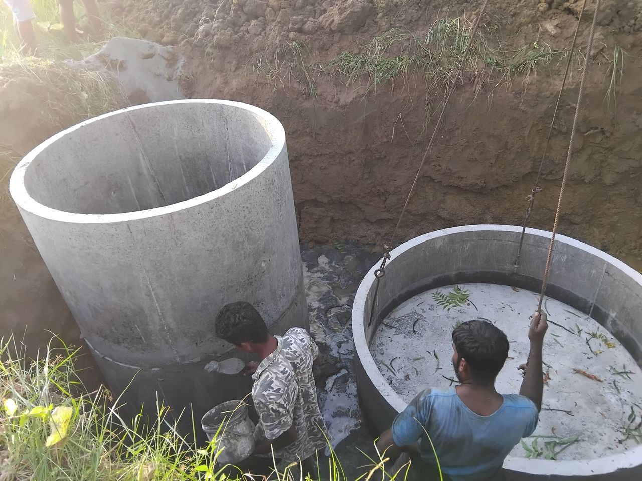 Two workers in a pit guiding a concrete ring on the crane rope beside a ring already in place