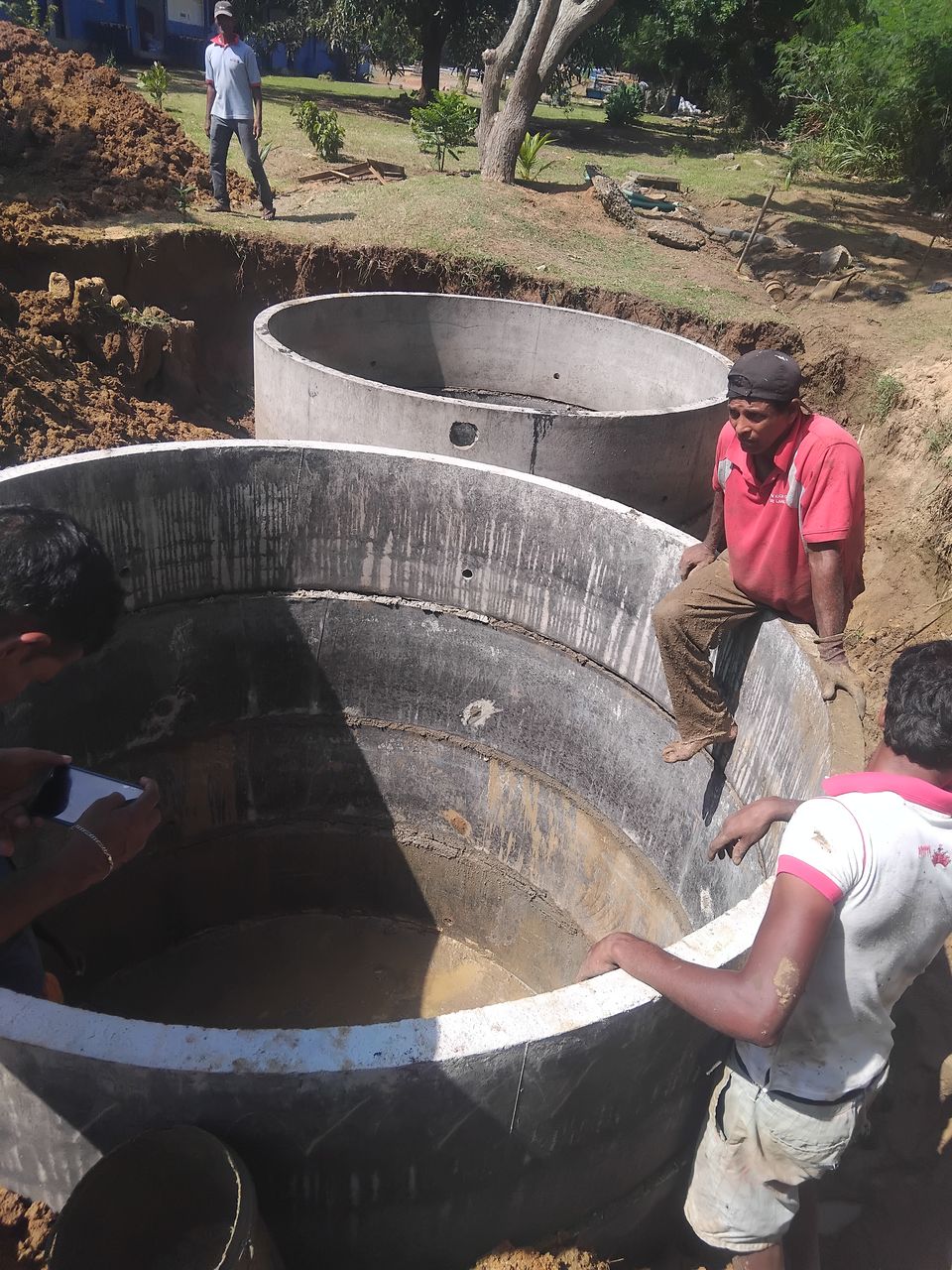 Workers around two large concrete rings set in a wide pit beside a lawn