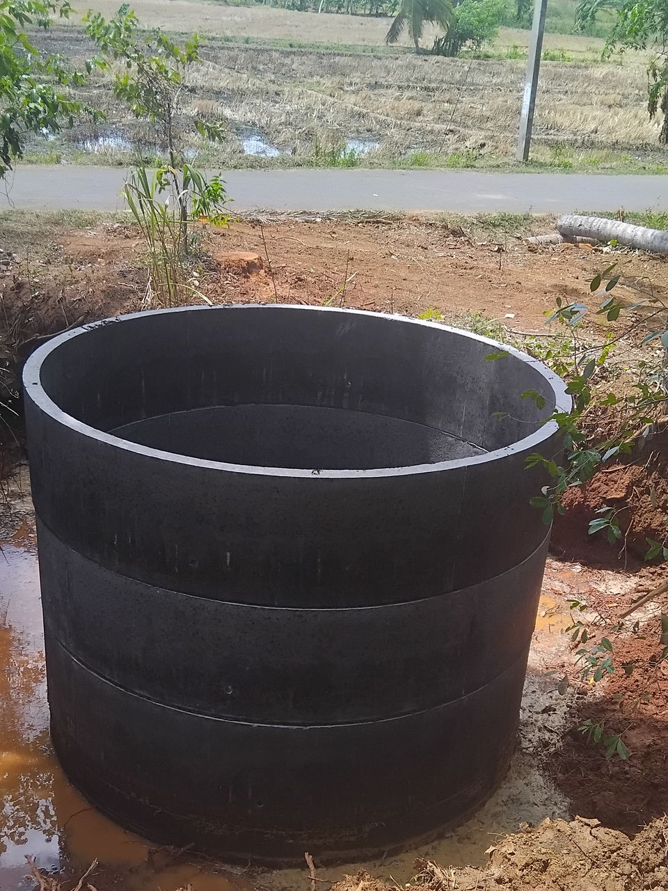 A new well lined with dark concrete rings, its top rings standing above the ground, with paddy fields behind and a water pipe laid to it