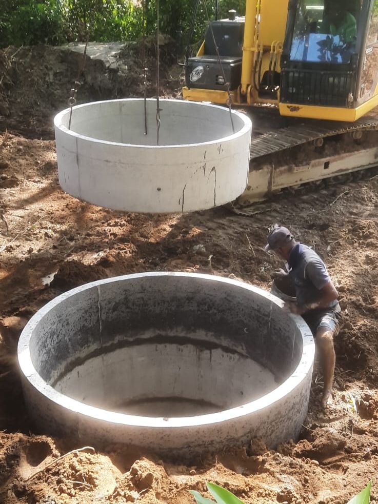 A concrete ring hanging from an excavator over a ring already set in the ground, a worker guiding it