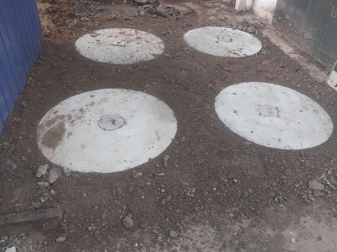 Four round concrete cover slabs set level with the ground in a passage beside a house, the chambers below them out of sight