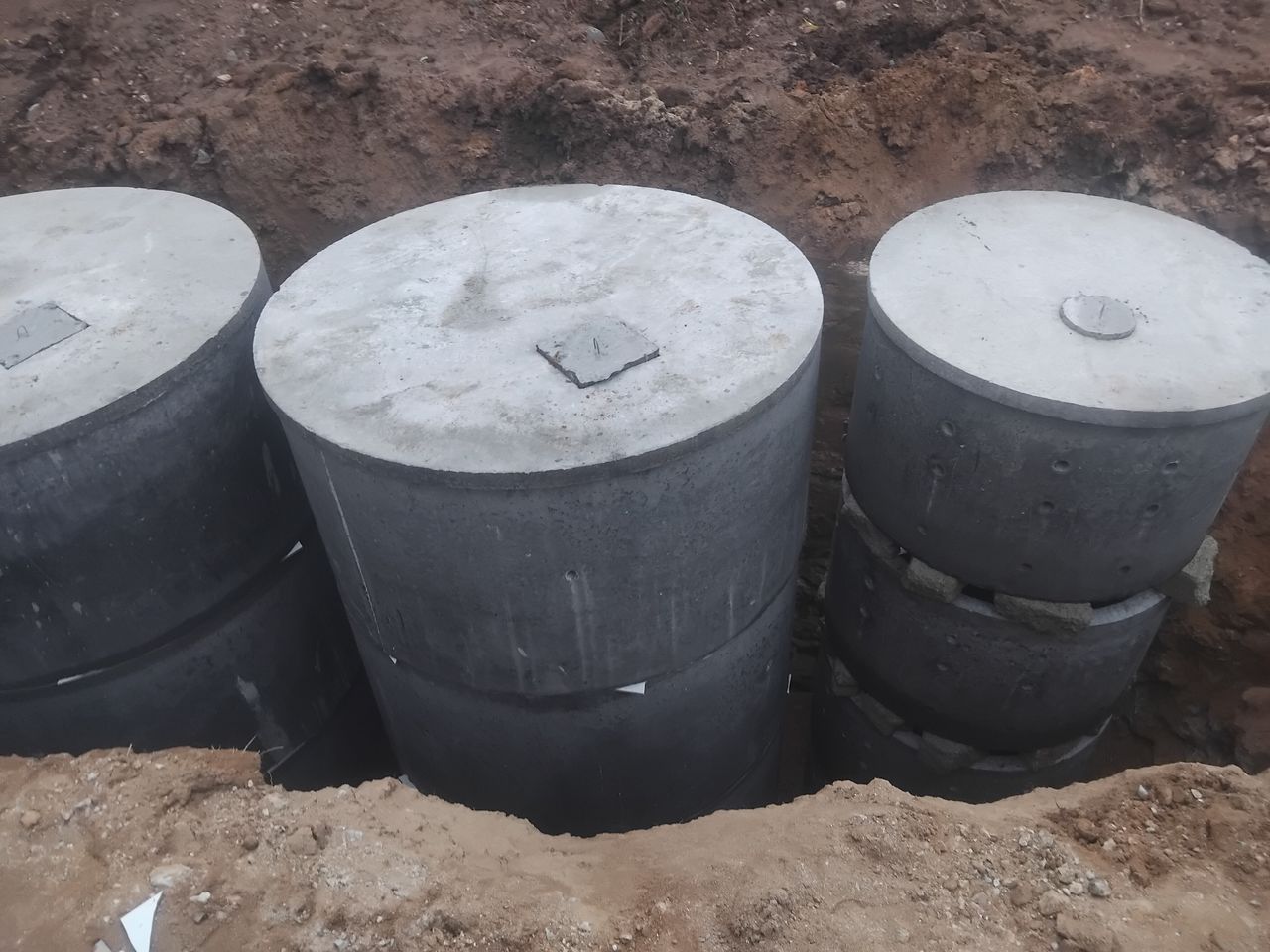 Two concrete ring chambers and a stack of perforated soakage rings in a pit, each with a cover slab and an access opening, before the ground is filled in