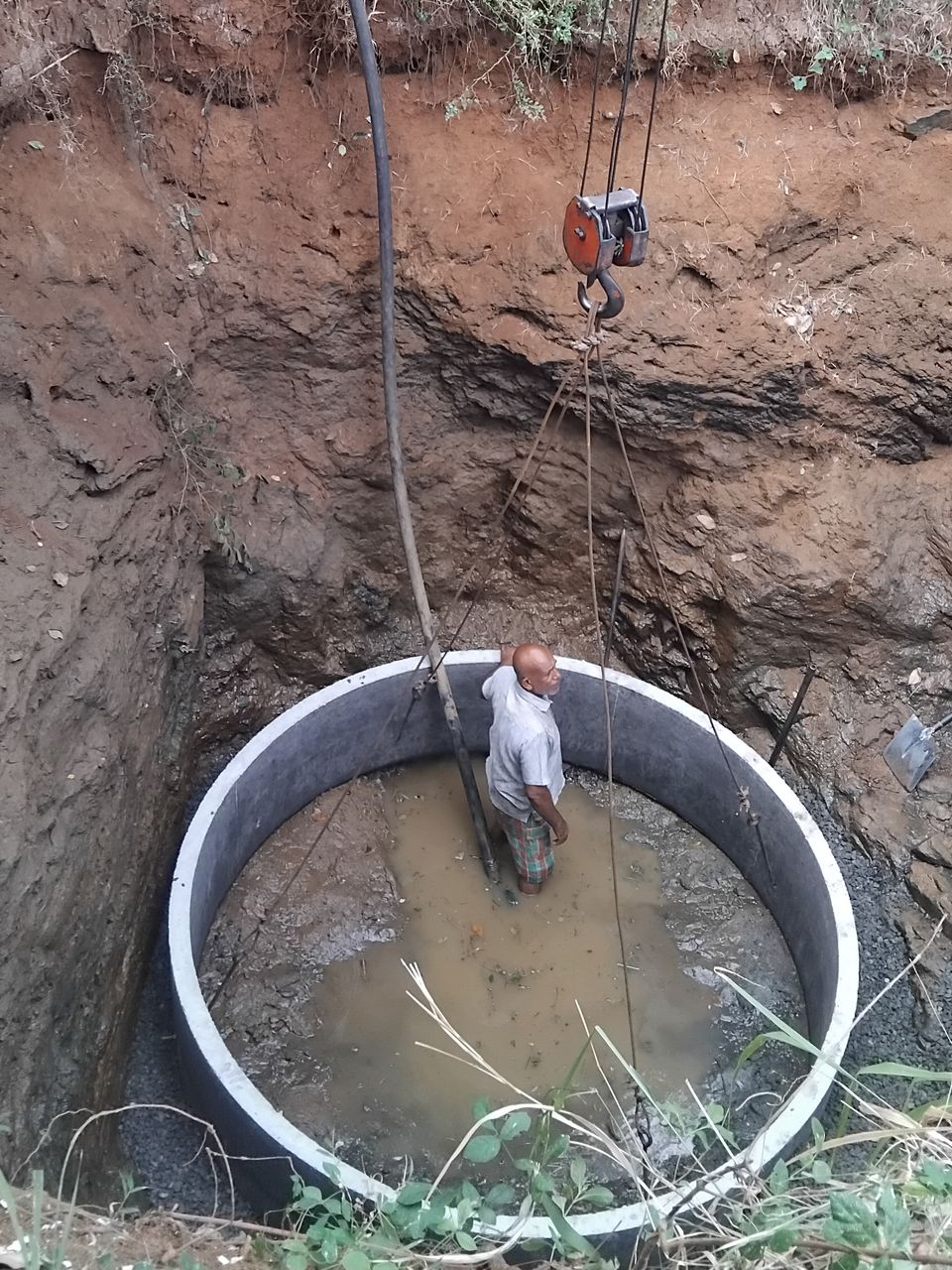 A concrete ring lowered by crane to the bottom of a deep well pit, standing in water, with a worker inside it